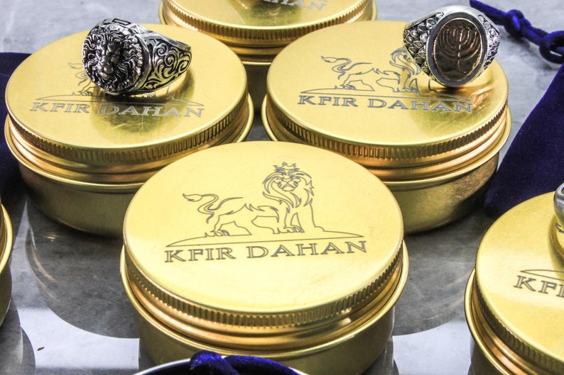 May include: Gold-colored metal boxes with a lion logo and the text "Kfir Dahan" engraved on the lid. The boxes are arranged on a white surface. A silver ring with a lion head design is on top of one of the boxes.