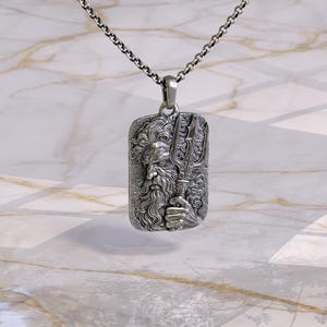 May include: A silver pendant necklace featuring a rectangular pendant with a detailed relief of a bearded figure holding a trident. The pendant is suspended from a silver chain. The design is intricate and the metal has a polished finish.