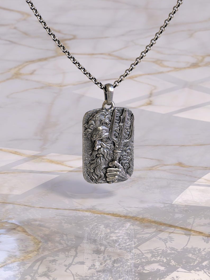 May include: A silver pendant necklace featuring a rectangular pendant with a detailed relief of a bearded figure holding a trident. The pendant is suspended from a silver chain. The design is intricate and the metal has a polished finish.