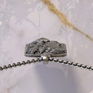 May include: A silver-toned pendant necklace featuring a rectangular pendant with a detailed relief design. The pendant is attached to a chain necklace. The background is a soft, blurred neutral tone.