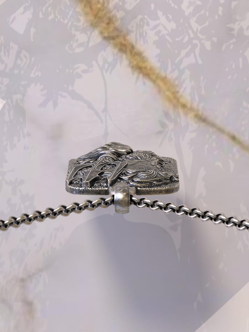 May include: A silver-toned pendant necklace featuring a rectangular pendant with a detailed relief design. The pendant is attached to a chain necklace. The background is a soft, blurred neutral tone.