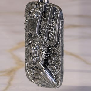 May include: A rectangular silver pendant featuring a detailed relief of a bearded man holding a trident. The pendant is suspended from a silver chain. The design has intricate details and a textured finish, suggesting a mythological theme.