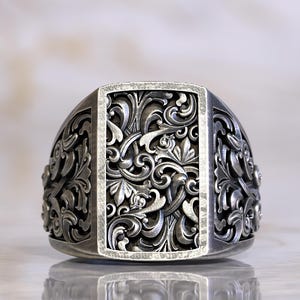 Könnte beinhalten: Ein silberfarbener Ring mit einem kunstvollen, floralen Design. Der rechteckige Mittelteil weist ein detailliertes Muster auf, das von einem erhabenen Rand umgeben ist. Die Seiten des Rings zeigen ebenfalls ein ähnliches, wirbelndes Motiv. Der Ring ist stark reflektierend.