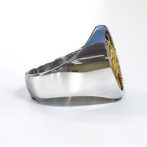 K&ouml;nnte beinhalten: Ein silberner Ring mit einem goldfarbenen Design auf der Oberseite. Der Ring ist oval geformt und hat eine glatte, polierte Oberfl&auml;che.