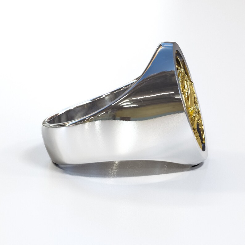 K&ouml;nnte beinhalten: Ein silberner Ring mit einem goldfarbenen Design auf der Oberseite. Der Ring ist oval geformt und hat eine glatte, polierte Oberfl&auml;che.