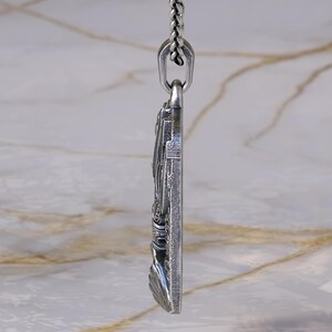 May include: A silver pendant with intricate detailing, suspended from a matching chain. The pendant features a detailed relief design, possibly religious or historical in theme. The chain is a classic link style, and the pendant is rectangular in shape.