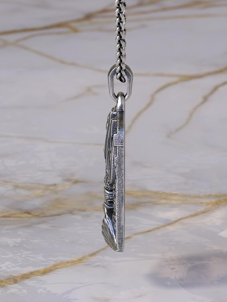May include: A silver pendant with intricate detailing, suspended from a matching chain. The pendant features a detailed relief design, possibly religious or historical in theme. The chain is a classic link style, and the pendant is rectangular in shape.