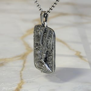 May include: A silver pendant necklace featuring a rectangular pendant with a detailed relief of a bearded figure holding a trident. The pendant is suspended from a delicate chain.