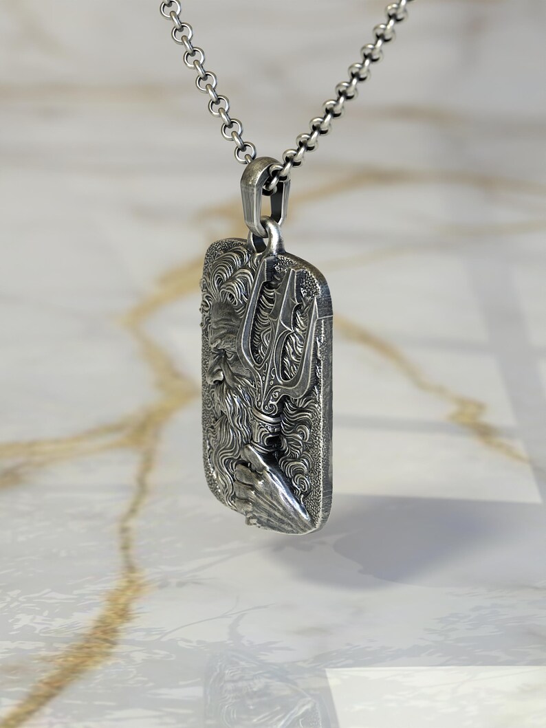 May include: A silver pendant necklace featuring a rectangular pendant with a detailed relief of a bearded figure holding a trident. The pendant is suspended from a delicate chain.