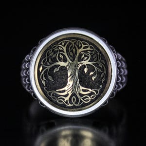 Puede incluir: Un anillo de plata con un diseño de árbol de la vida negro y dorado. El anillo tiene una banda texturizada.