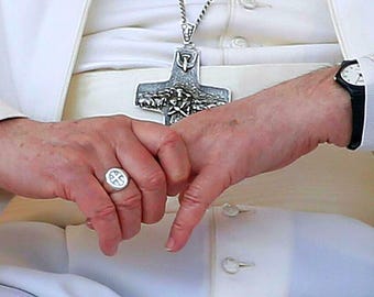 May include: A close-up of a person's hands wearing a silver ring with a cross design. The person is wearing a white robe and a silver chain with a cross pendant.