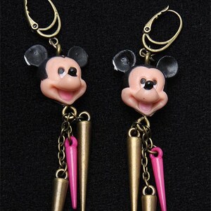 May include: A pair of gold-tone earrings featuring a black and white Mickey Mouse head charm with pink accents. Each earring has a long, gold-tone spike with a pink accent.