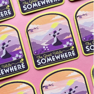 May include: A set of embroidered patches featuring a purple landscape with a woman in a purple dress dancing in a field of flowers. The patches have a yellow border and the text "The Great Wide Somewhere" in black.