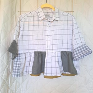 May include: A white and grey checked shirt with grey and white gingham detailing on the sleeves and a grey frill at the waist. The shirt is button-down and hangs on a yellow hanger.