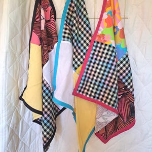 May include: Three patterned fabric squares hang from a gold wire grid. The squares feature floral, gingham, and abstract designs. Each square has a different colour border, including black, pink, and blue. The squares are made of fabric.