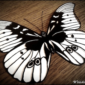 May include: A black and white paper cut-out of a butterfly with intricate details. The butterfly is set against a wooden background.