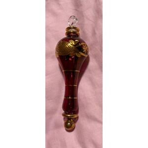 May include: A deep red glass finial ornament featuring a teardrop shape with gold-painted accents. The top includes a clear glass bead finial, while the body is decorated with gold horizontal bands and etched floral patterns in a metallic gold finish.