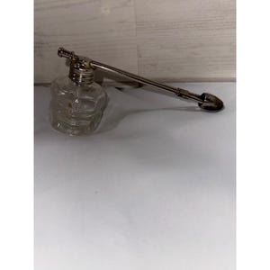 May include: A clear glass perfume bottle with a silver metal sprayer. The bottle has a unique, faceted design. The sprayer extends from the bottle with a long, straight tube ending in a small, rounded nozzle. The bottle sits on a white surface.