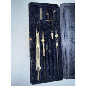 May include: A vintage drafting instrument set housed in a rectangular case with a dark blue velvet interior. The set includes four metal drawing tools, including a compass and various precision pens with silver and black components.