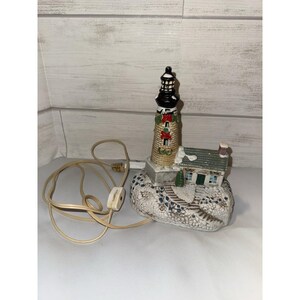 May include: A ceramic lighthouse lamp with a black top and a beige brick tower, adorned with a festive wreath. The lamp includes a small house with a snow-covered roof and a winding path. The lamp is connected to a power cord.