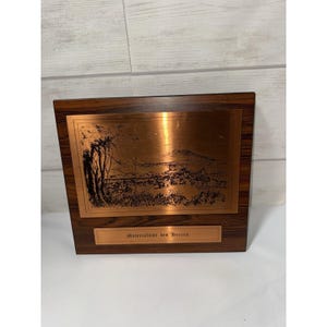 May include: A rectangular copper plaque with a landscape scene, set within a dark wood frame. The plaque features a detailed etching of trees, a town, and mountains. The text "Materialamt des Heeres" is inscribed on a smaller copper plate below the landscape.
