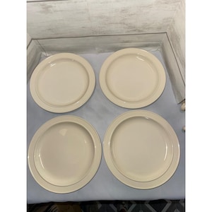 May include: A set of four round, cream-colored ceramic dinner plates arranged in a two-by-two grid. Each plate features a smooth, solid-colored surface with a slightly raised, wide rim that frames the central area of the plate.