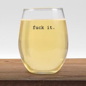 Puede incluir: Una copa de vino sin tallo llena de un líquido amarillo claro. Las palabras "fuck it." están impresas en negro en el vaso. El vaso está sobre una superficie de madera.