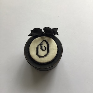 May include: A small, round, black box with a white fabric top featuring a black embroidered letter "G". The box has a black ribbon tied around it.