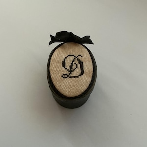 May include: A small, round, dark-colored box with a lid featuring a beige fabric insert. The insert is embroidered with a black cross-stitch letter D in a decorative font. A small black ribbon bow is attached to the top edge of the lid.