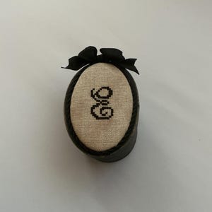 May include: An oval-shaped pincushion featuring a beige linen top with the black cross-stitched letters "G&G" in the center. The cushion is encased in a dark brown frame with a decorative black twisted cord trim and a black fabric bow on top.