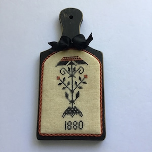 May include: A black wooden cutting board with a cream-colored fabric panel featuring a black embroidered design of a stylized tree with the year 1880 stitched below. The board has a black ribbon tied around the top.