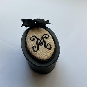 May include: An oval black box featuring a beige fabric insert on the lid with a black cross-stitched letter M. The lid is decorated with a black twisted rope border and a black fabric bow attached to the top edge of the oval frame.
