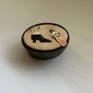 May include: A black cross-stitch design of a high-heeled shoe on a tan fabric, set in a round, black container. The container has a small wooden tag with the number "No. 489" attached. Several pins are inserted into the fabric.