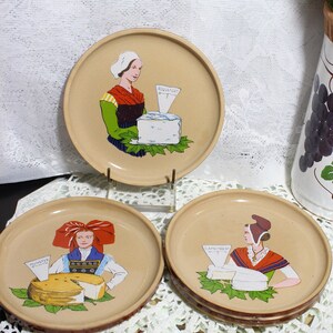 May include: Three ceramic plates with brown rims feature illustrations of women in traditional clothing holding cheese wedges. The plates are labeled "Munster", "Camembert", and "Roquefort".