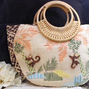 May include: A woven straw handbag with a bamboo handle. The bag features embroidered palm trees, monkeys, and foliage in shades of brown, green, yellow, and orange. The bag's edges are trimmed with a brown and tan woven pattern.