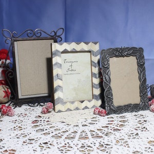 May include: Three decorative picture frames in various styles. One is a wrought iron design, another has a chevron pattern, and the third features a floral motif. The middle frame has text that reads "Treasures of India, handcrafted in India."