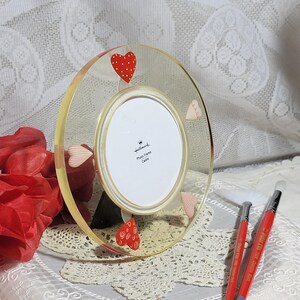 May include: A clear acrylic photo frame with a gold trim and a white mat. The frame is decorated with red and white hearts. The frame is sitting on a white lace tablecloth. The text on the frame reads "Hallmark Photo Frame Cadre".