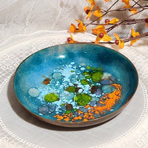 May include: A round, shallow bowl with a turquoise interior and a bronze-colored rim. The interior features a colorful abstract design with blue, green, and orange accents. The bowl is set against a white lace background with orange floral accents.