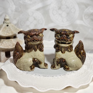May include: Two ceramic foo dog figurines with brown and white glaze. The figurines are sitting on a white lace doily.