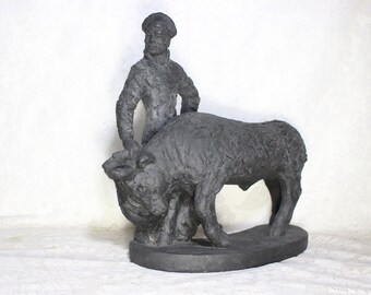 May include: A dark gray sculpture depicting a figure standing next to a bull. The figure is wearing a hat and coat, with hands on hips. The bull is large and muscular, with a textured surface. The sculpture is on a dark base.