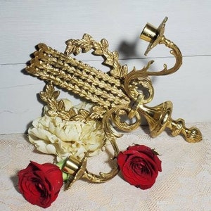 May include: Ornate gold-colored wall sconce with two candle holders, featuring a decorative leaf and scroll design. Two red roses and a cream-colored flower are placed near the sconce. The background is a white wooden surface.