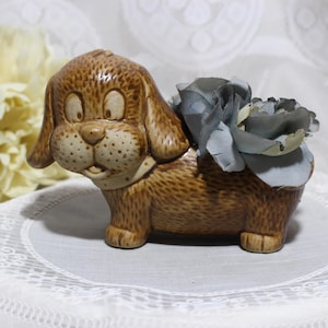 May include: A brown ceramic dog figurine with a light brown and white flower on its back. The dog has a brown nose and black eyes.