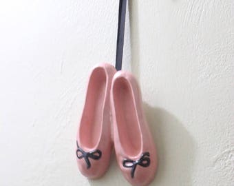May include: A pair of pink ballet slippers with black bows, hanging from a black ribbon. The slippers are a soft pink color and are hanging against a white wall.