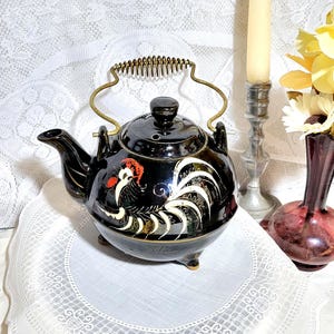 May include: Black ceramic teapot with a rooster design and a spring-loaded handle. The teapot has three feet and is sitting on a white lace doily.
