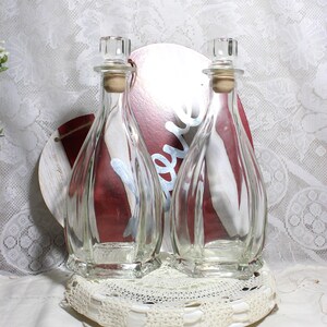 May include: Two clear glass bottles with cork stoppers and clear glass tops. The bottles sit on a white lace doily. A red and white decorative piece is behind the bottles.