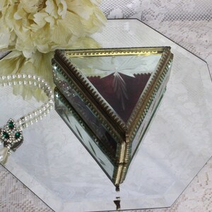 May include: A triangular glass jewelry box with a gold-colored metal frame. The box has a starburst design etched into the glass and a burgundy interior. A pearl necklace with a green gem pendant and a white flower are also in the image.