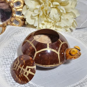 May include: A small, brown coconut shell turtle trinket box with a gold painted design. The box has a lid and a small, yellow eye on the side.