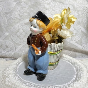 May include: A ceramic figurine of a boy wearing a black hat and brown jacket, holding a pipe. He is standing next to a planter with yellow and white flowers.