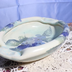 May include: Oval-shaped ceramic ashtray with a white base and blue floral design. The rim is painted with blue and green accents. The ashtray is placed on a white lace doily.