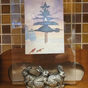 May include: A silver-toned metal figurine of three rabbits sitting together. The rabbits are in a picture frame with a watercolor painting of a tree with birds perched on the branches. The painting has a light blue, pink, and white color scheme.